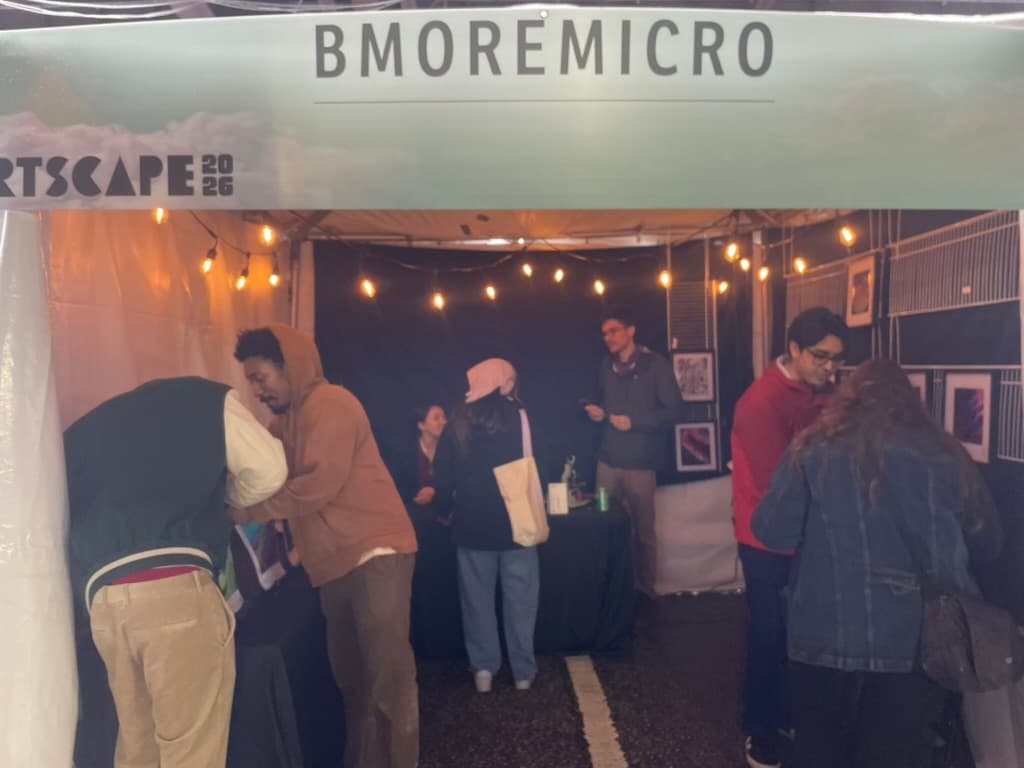 Visitors inside the studio's tent at Artscape 2026, under a banner reading “Bmoremicro” and string lights, with framed prints on black walls.
