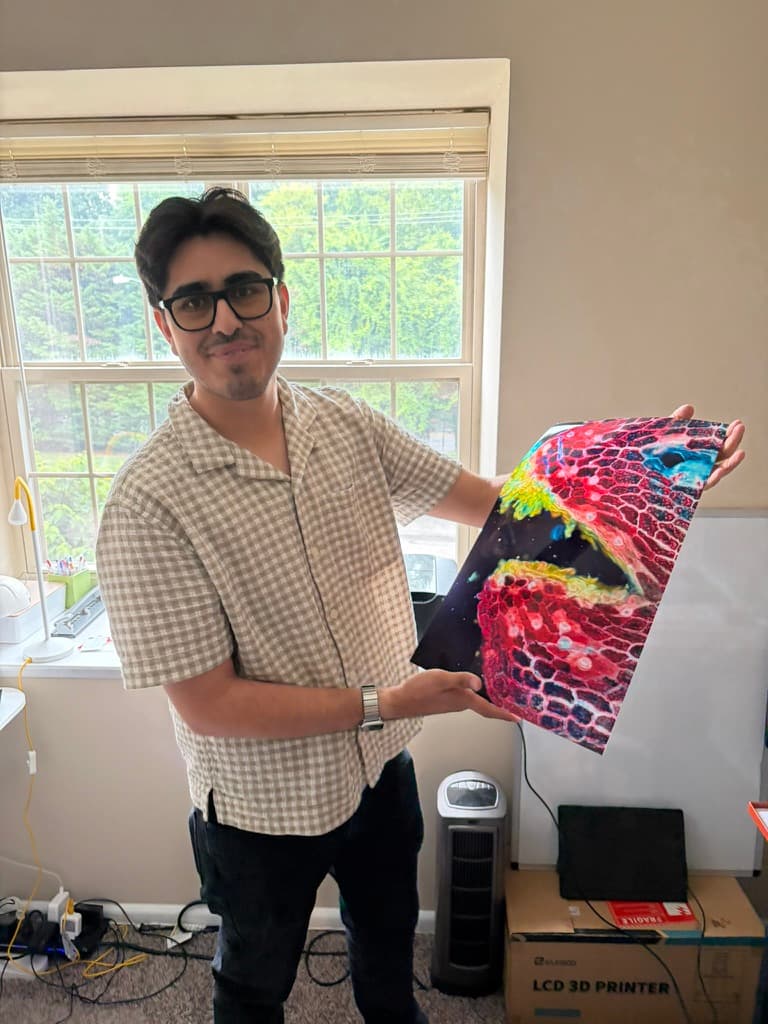 One of the studio's makers at home by a window, holding up a large unframed print of a micrograph in reds, blues and yellow-green.