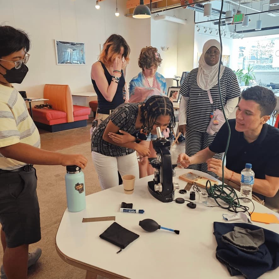 A student leans in to look through a black microscope on a white table while other students watch and one of the studio's makers adjusts the slide.