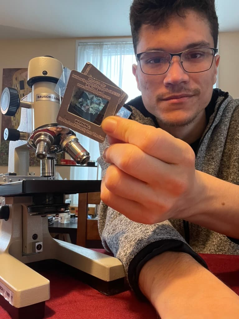 One of the studio's makers holding a mounted glass slide up to the camera beside a cream Bausch & Lomb microscope, its objectives catching the light.