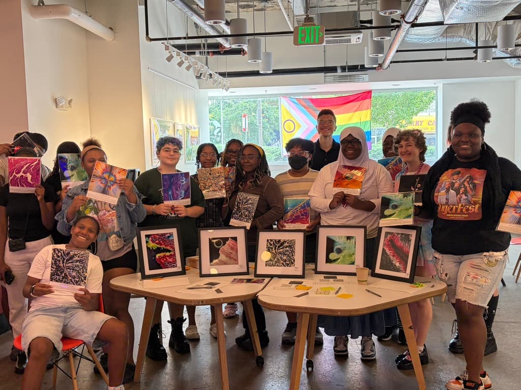 A group of students in a bright gallery space, smiling and holding up micrograph prints, with five framed prints standing on the tables in front of them.